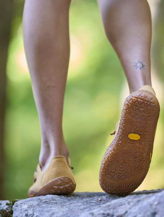 Vista dal basso delle gambe di una persona che cammina su una roccia all’aperto, indossando scarpe minimal color sabbia con suola in gomma, in un ambiente naturale verde.