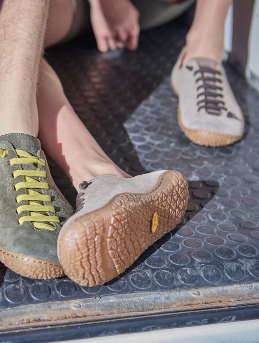 Primo piano di più persone sedute su una superficie metallica, con scarpe minimal in diversi colori; una suola in gomma flessibile è ben visibile in primo piano.